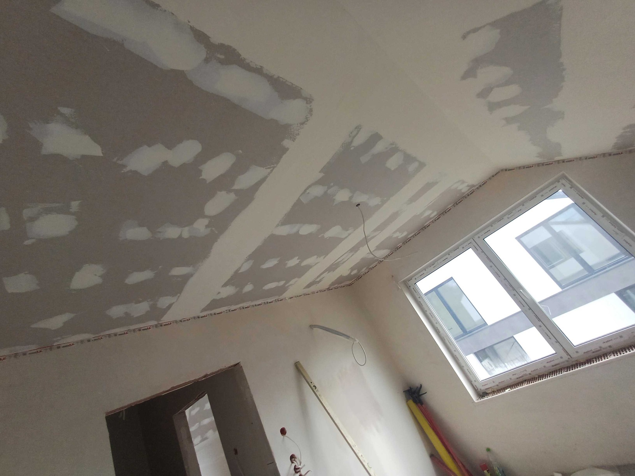 Pokój w trakcie gładzenia sufitu, widoczne ślady po łatach gipsowych, okno dachowe i narzędzia malarskie przy ścianie. Prace wykończeniowe w nowym budownictwie.