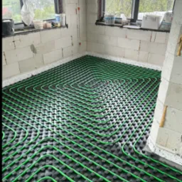 Zielone rury ogrzewania podłogowego ułożone spiralnie na czarnej macie w surowym wnętrzu, widok z góry na instalację przed zalaniem posadzką.