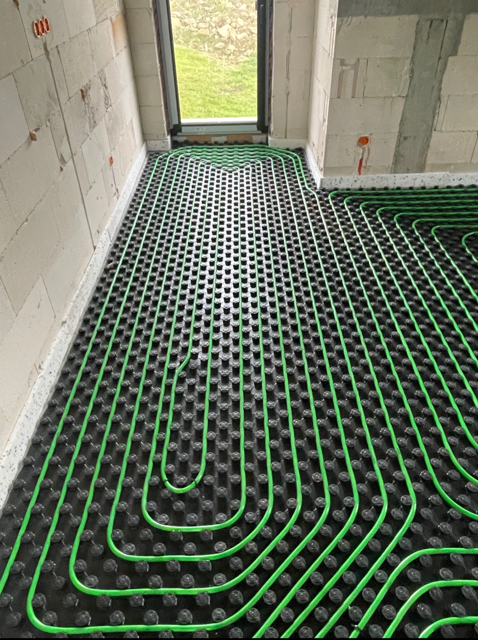 Zielone rury ogrzewania podłogowego ułożone na czarnej macie w surowym wnętrzu. Widoczne fragmenty ścian z białego pustaka i okno z widokiem na zewnątrz.