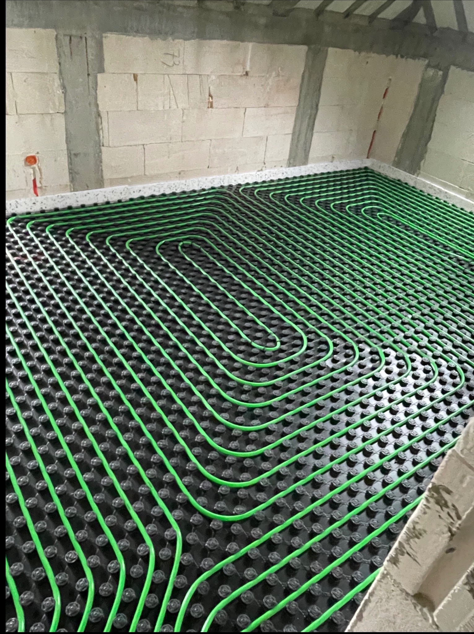 Ułożone zielone rury ogrzewania podłogowego na czarnej macie izolacyjnej, widoczne ściany z betonu komórkowego w surowym stanie.
