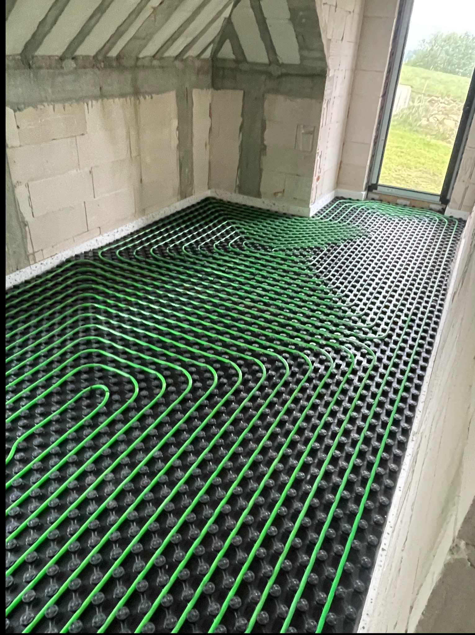 Zielone rury ogrzewania podłogowego ułożone na czarnej macie w surowym wnętrzu z oknem. Instalacja przed wylaniem posadzki, widok z góry.
