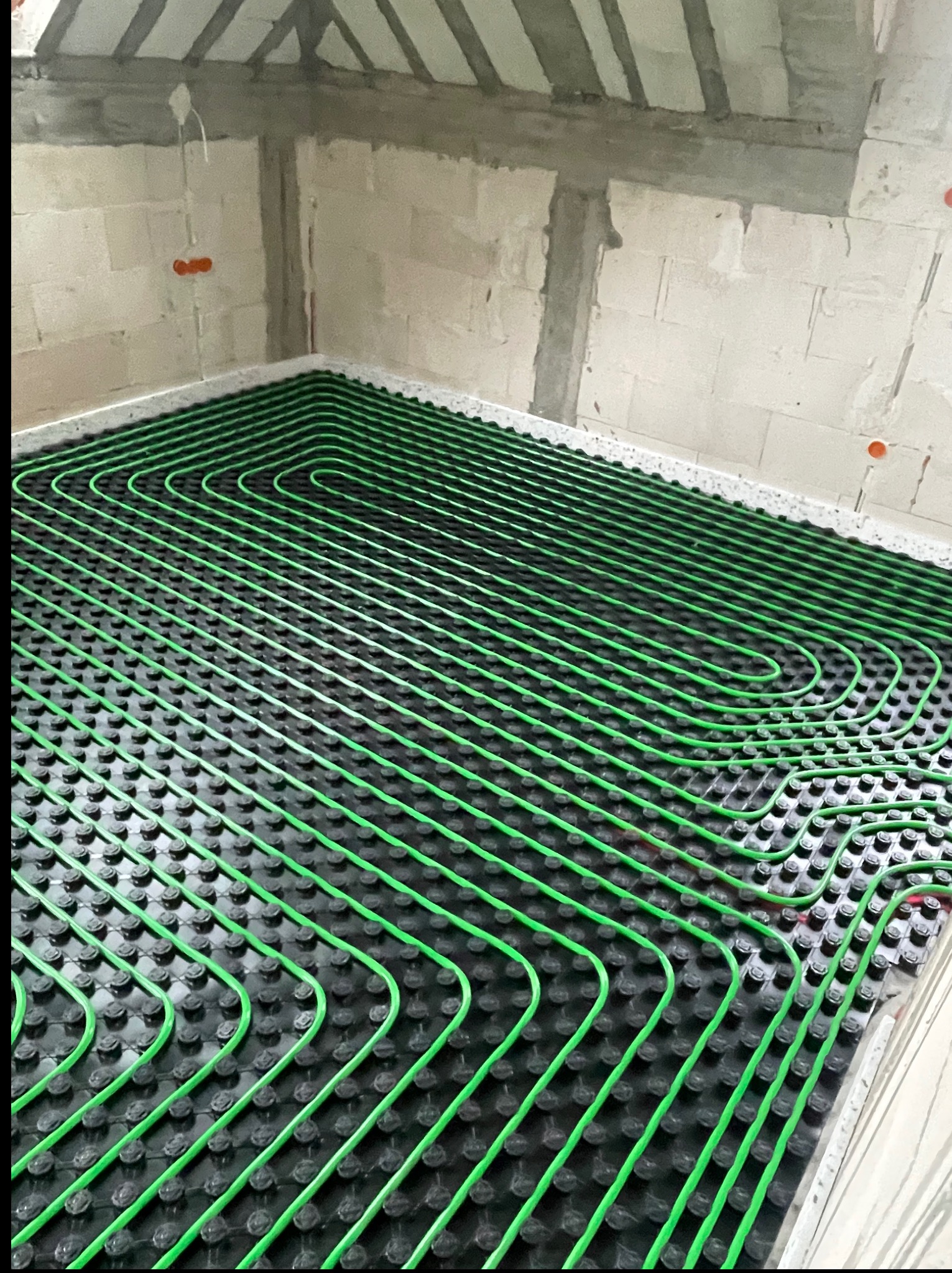 Zielone rury ogrzewania podłogowego na czarnej macie, ułożone w regularnych pętlach w surowym wnętrzu. Widoczne ściany z betonu komórkowego i fragment stropu.