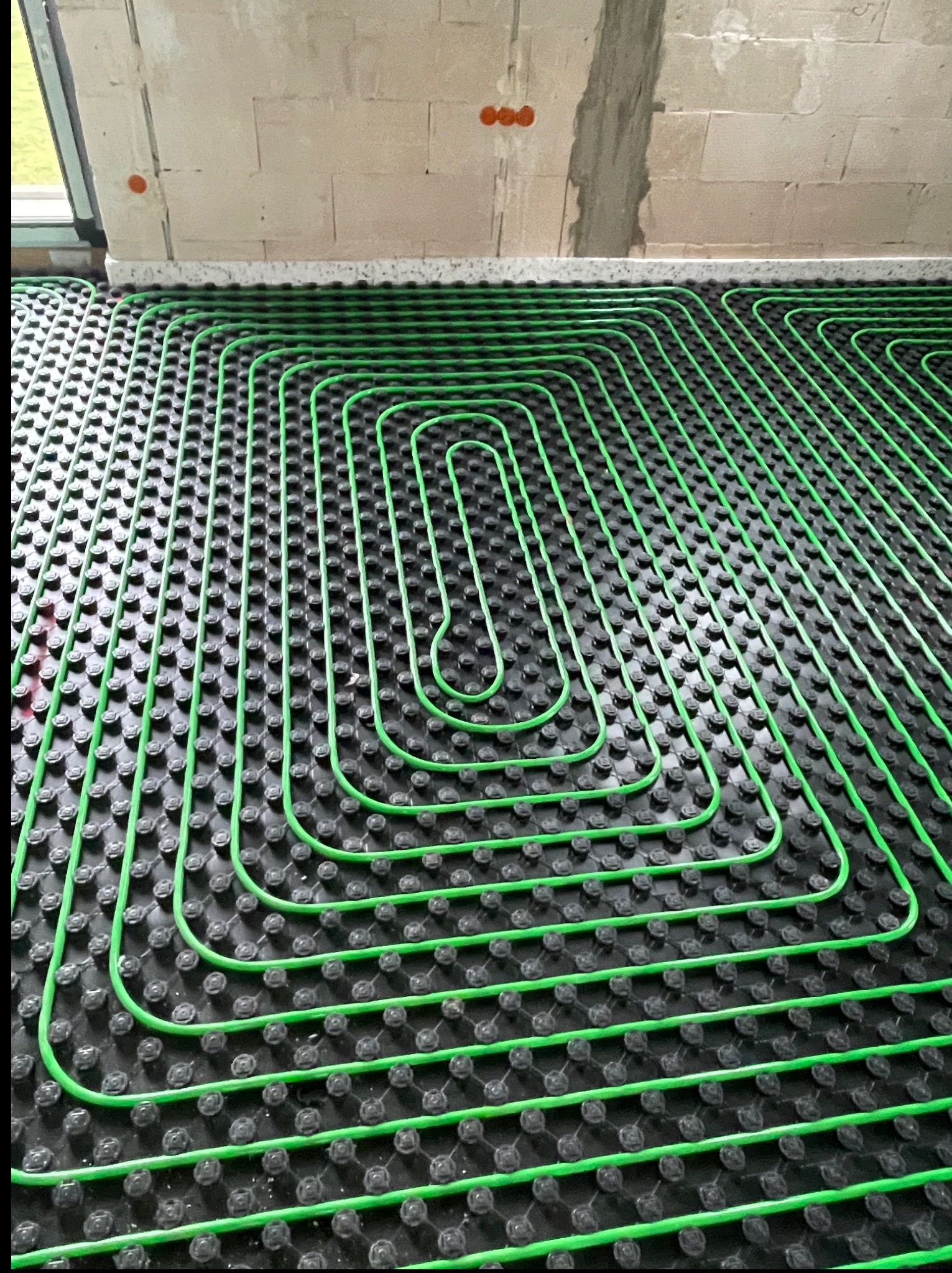 Zielone rury ogrzewania podłogowego ułożone na czarnej macie w kształt meandryczny, widoczne surowe ściany budynku w tle. Precyzyjny montaż instalacji grzewczej.
