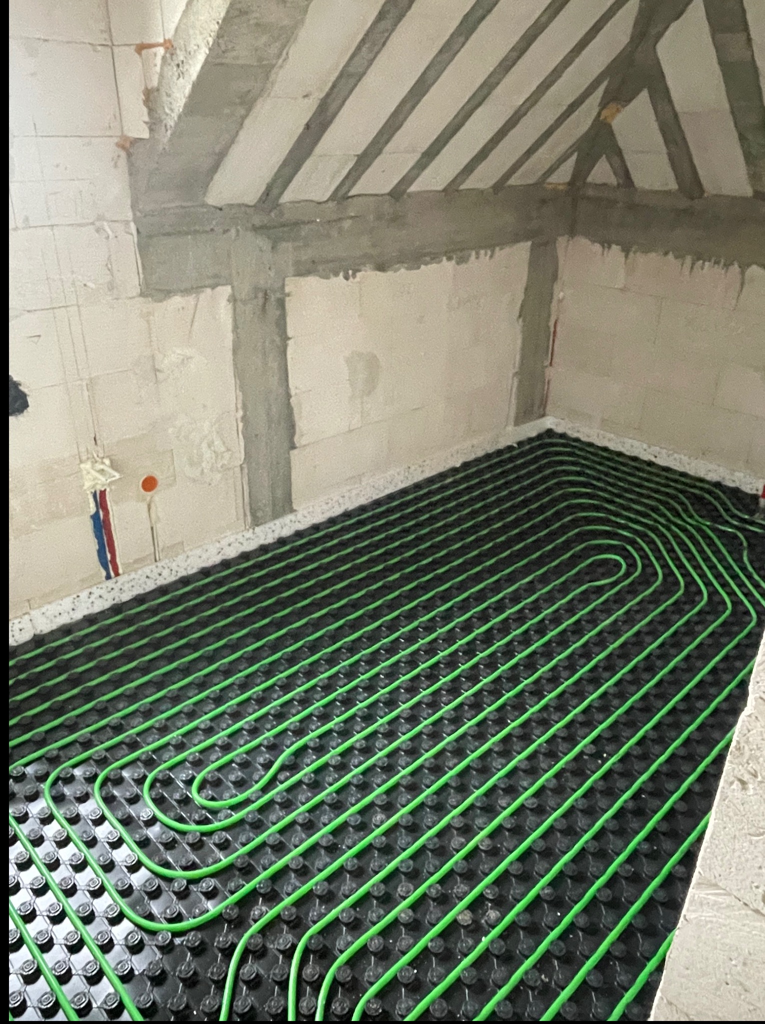 Zielone rurki ogrzewania podłogowego ułożone na czarnej macie w surowym wnętrzu z betonowym stropem. Widoczne pionowe przewody instalacyjne.
