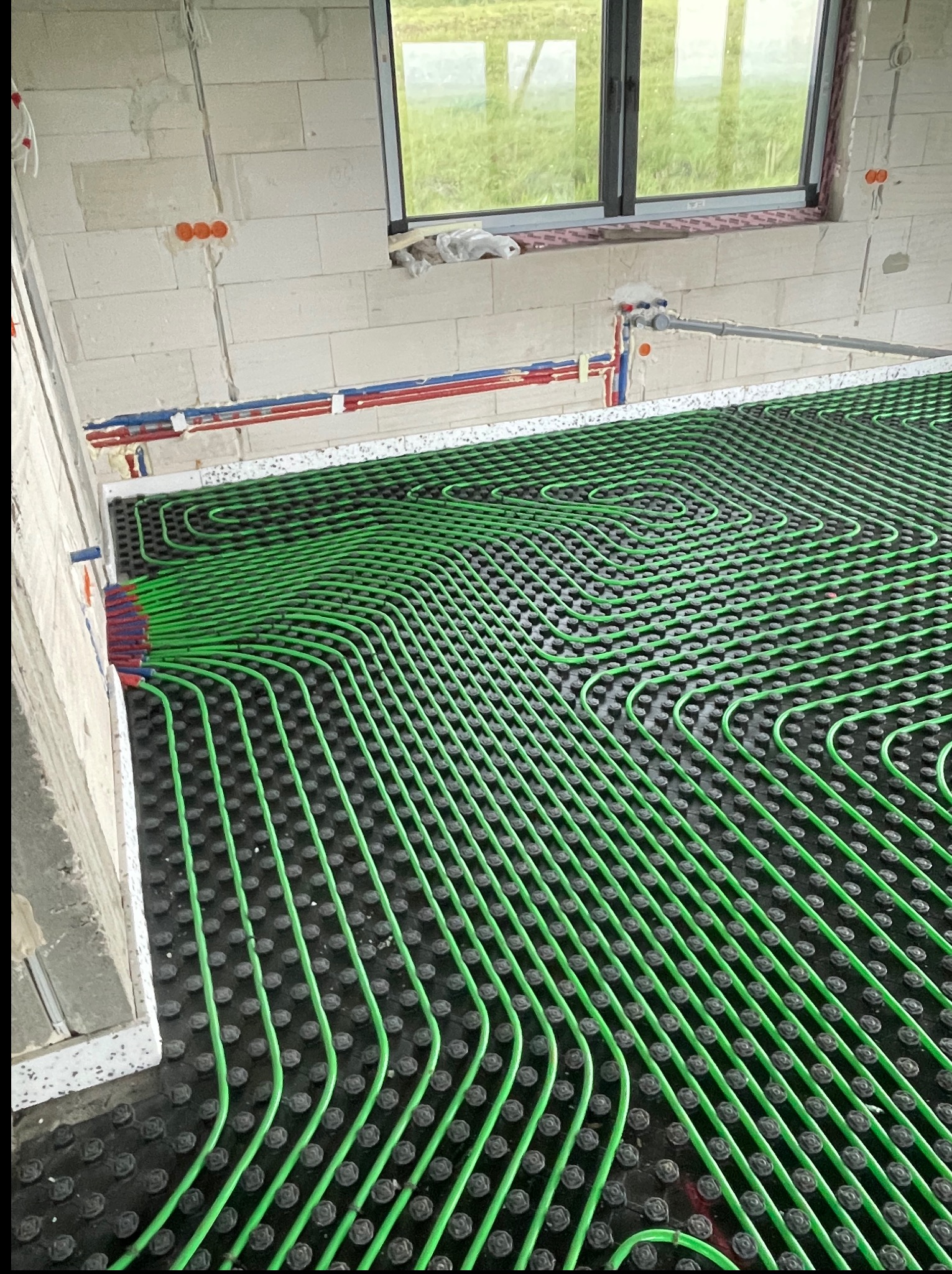 Zielone rury ogrzewania podłogowego ułożone na czarnej macie w surowym wnętrzu z oknem. Widoczne instalacje wodne na ścianie.