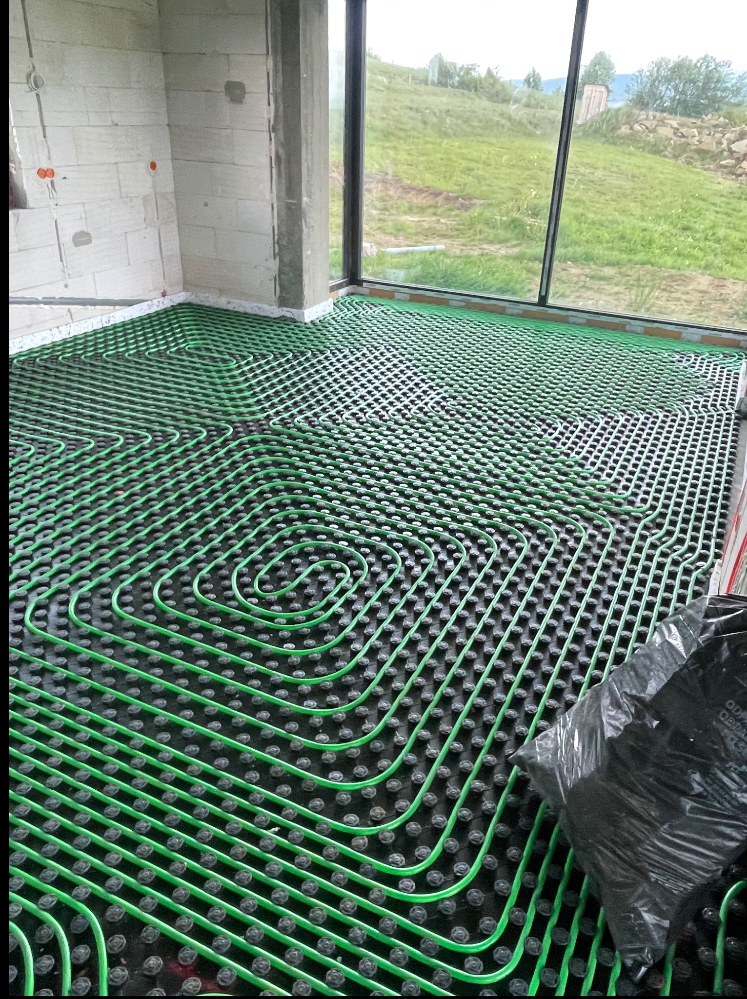 Zielone rury ogrzewania podłogowego ułożone spiralnie na czarnej macie w surowym wnętrzu z dużym oknem i widokiem na zieleń. Etap instalacji przed wylewką.