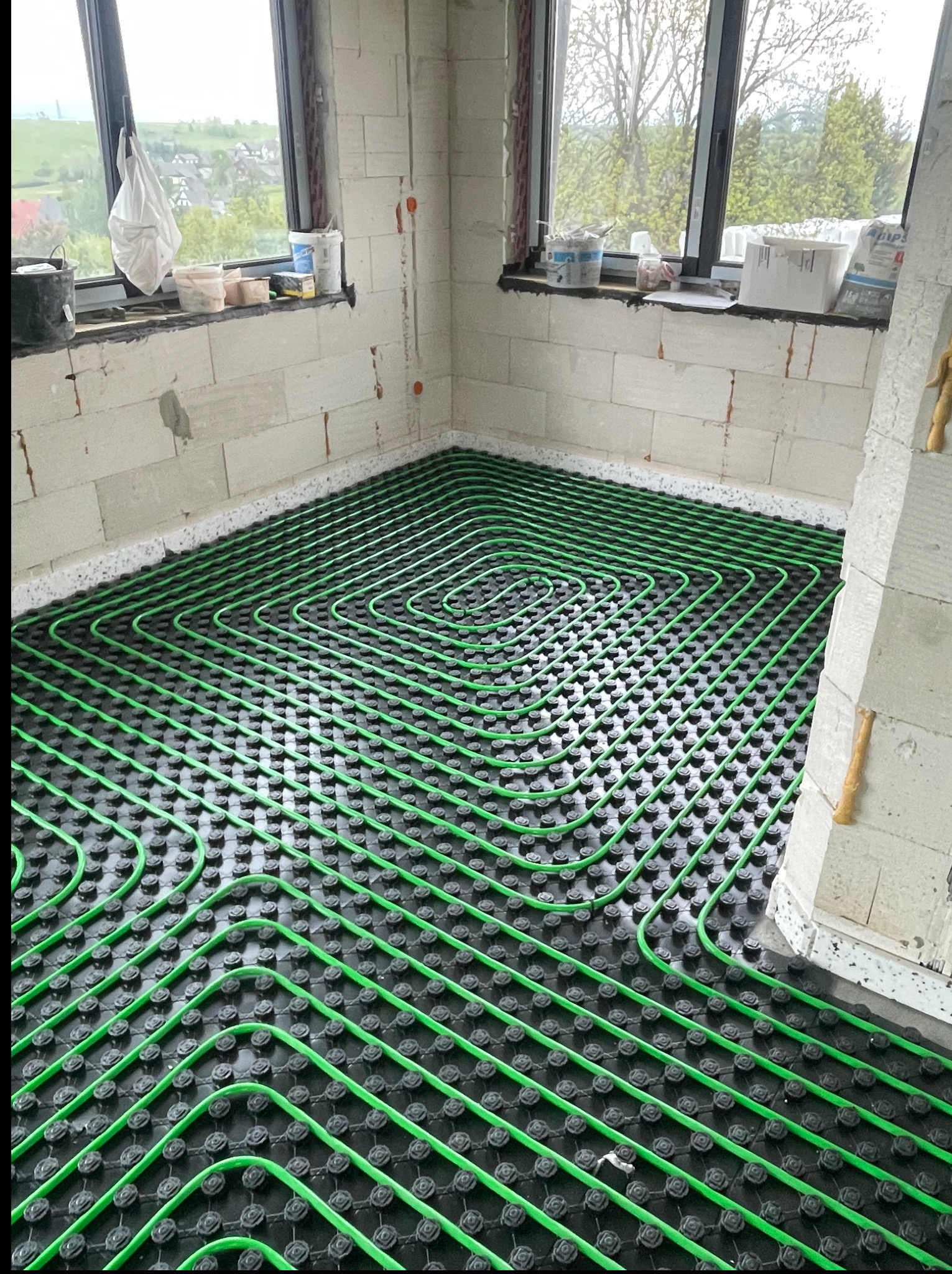 Zielone rury ogrzewania podłogowego ułożone spiralnie na czarnej macie w surowym wnętrzu, widok z góry na instalację przed zalaniem posadzką.