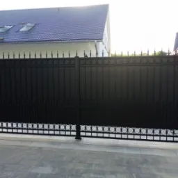 Czarna, metalowa brama wjazdowa z dekoracyjnymi elementami na tle jasnego domu z ciemnym dachem i brukowanym podjazdem. Brama osadzona na ceglanych słupkach.