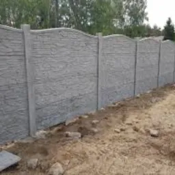 Szary, teksturowany płot z betonu imitującego kamień, z lekko falistym wykończeniem górnej krawędzi, na tle zieleni drzew i krzewów. Widoczne elementy montażowe.