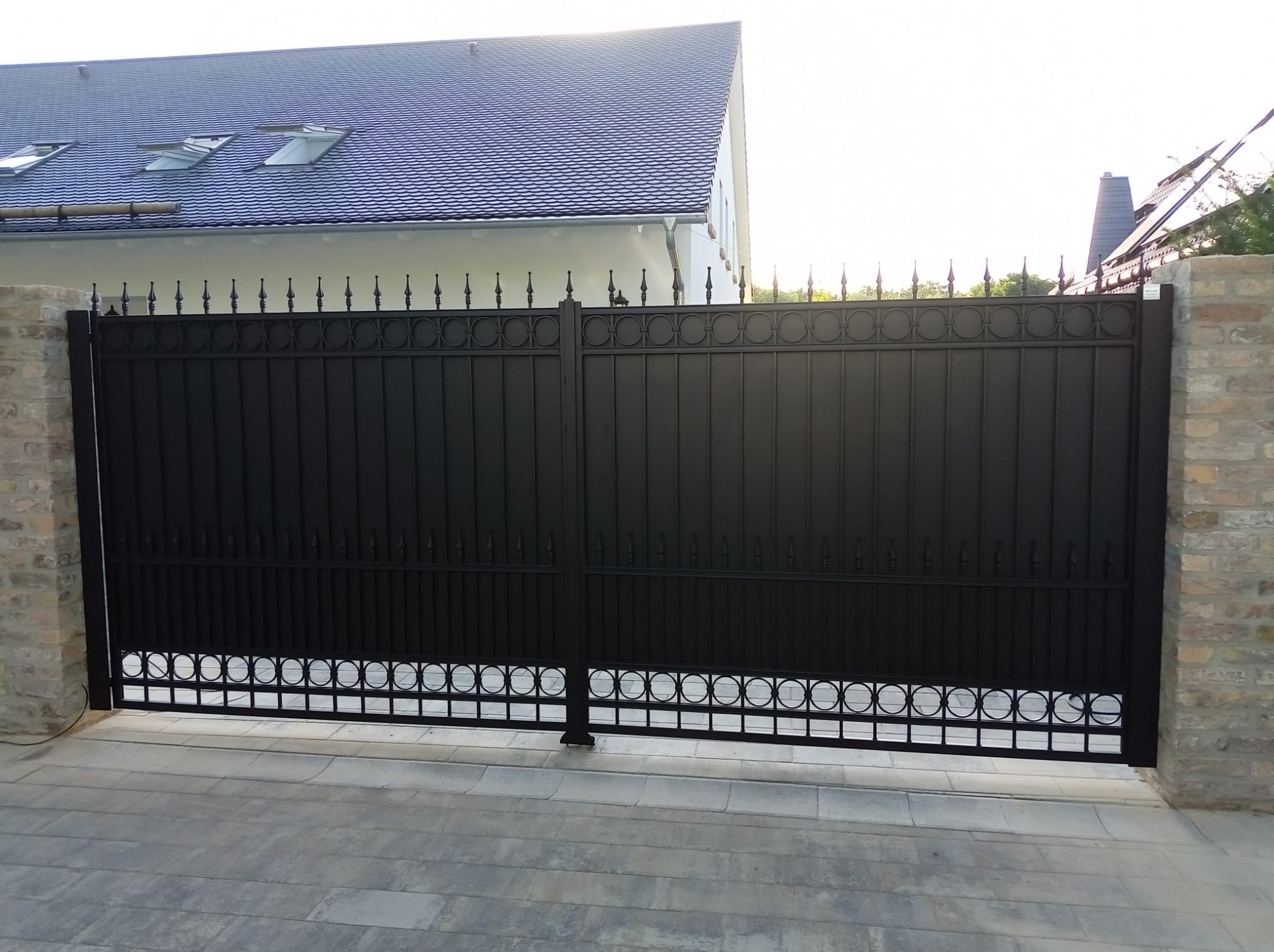 Czarna, metalowa brama wjazdowa z dekoracyjnymi elementami na tle jasnego domu z ciemnym dachem i brukowanym podjazdem. Brama osadzona na ceglanych słupkach.