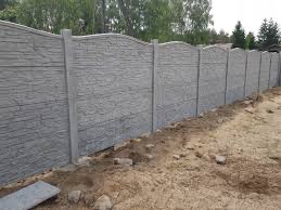 Szary, teksturowany płot z betonu imitującego kamień, z lekko falistym wykończeniem górnej krawędzi, na tle zieleni drzew i krzewów. Widoczne elementy montażowe.