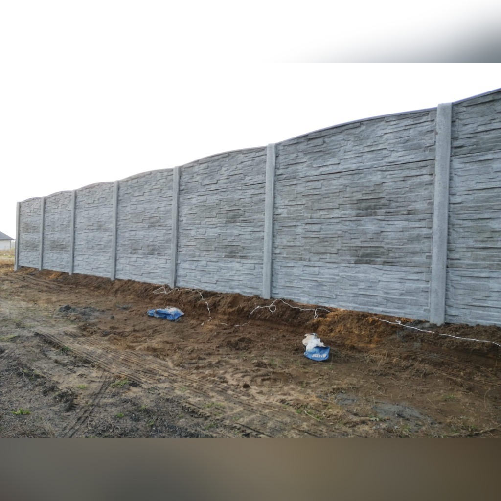 Długi, szary mur z betonowych elementów imitujących kamień, tworzący ogrodzenie na nierównym terenie. Widoczne ślady opon i resztki po montażu.