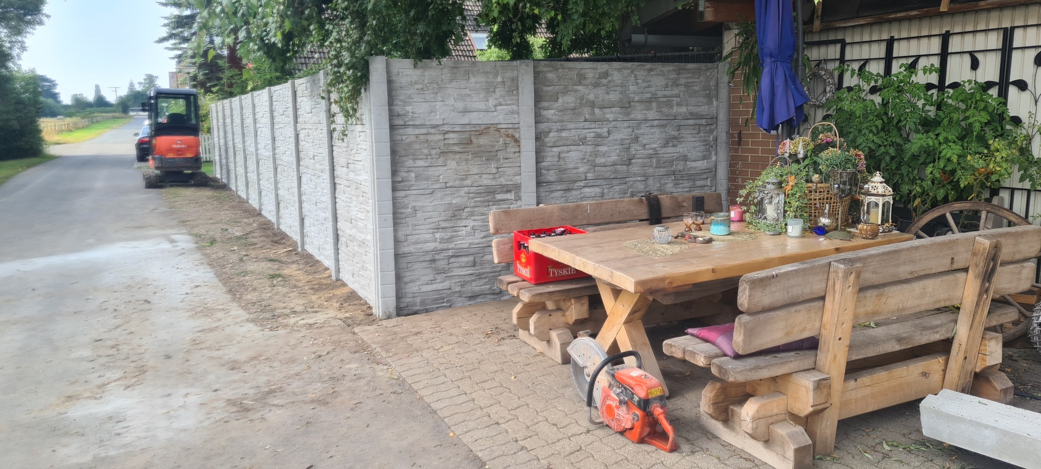 Nowo postawione ogrodzenie panelowe z betonu o fakturze kamienia. W tle mała koparka i stół z ławkami. Na pierwszym planie piła do betonu.