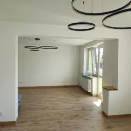 Jasne, przestronne wnętrze po remoncie z drewnianą podłogą i nowoczesnym, okrągłym oświetleniem. Widok na okno i przyległe pomieszczenie.