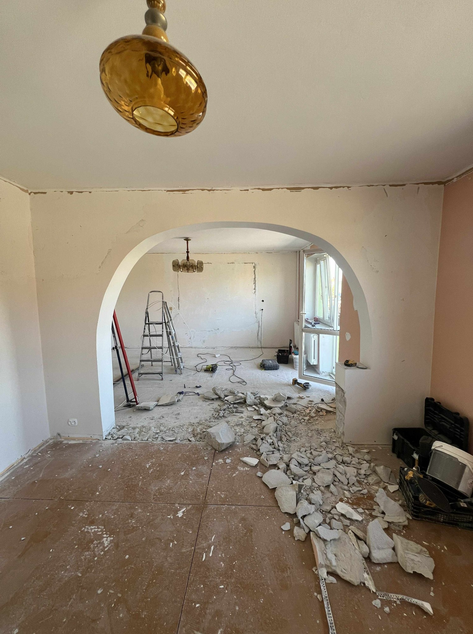 Demontaż ściany działowej z cegły w mieszkaniu. Gruz na podłodze, widoczne narzędzia i drabina. Prace remontowe w toku, surowy stan wnętrza.