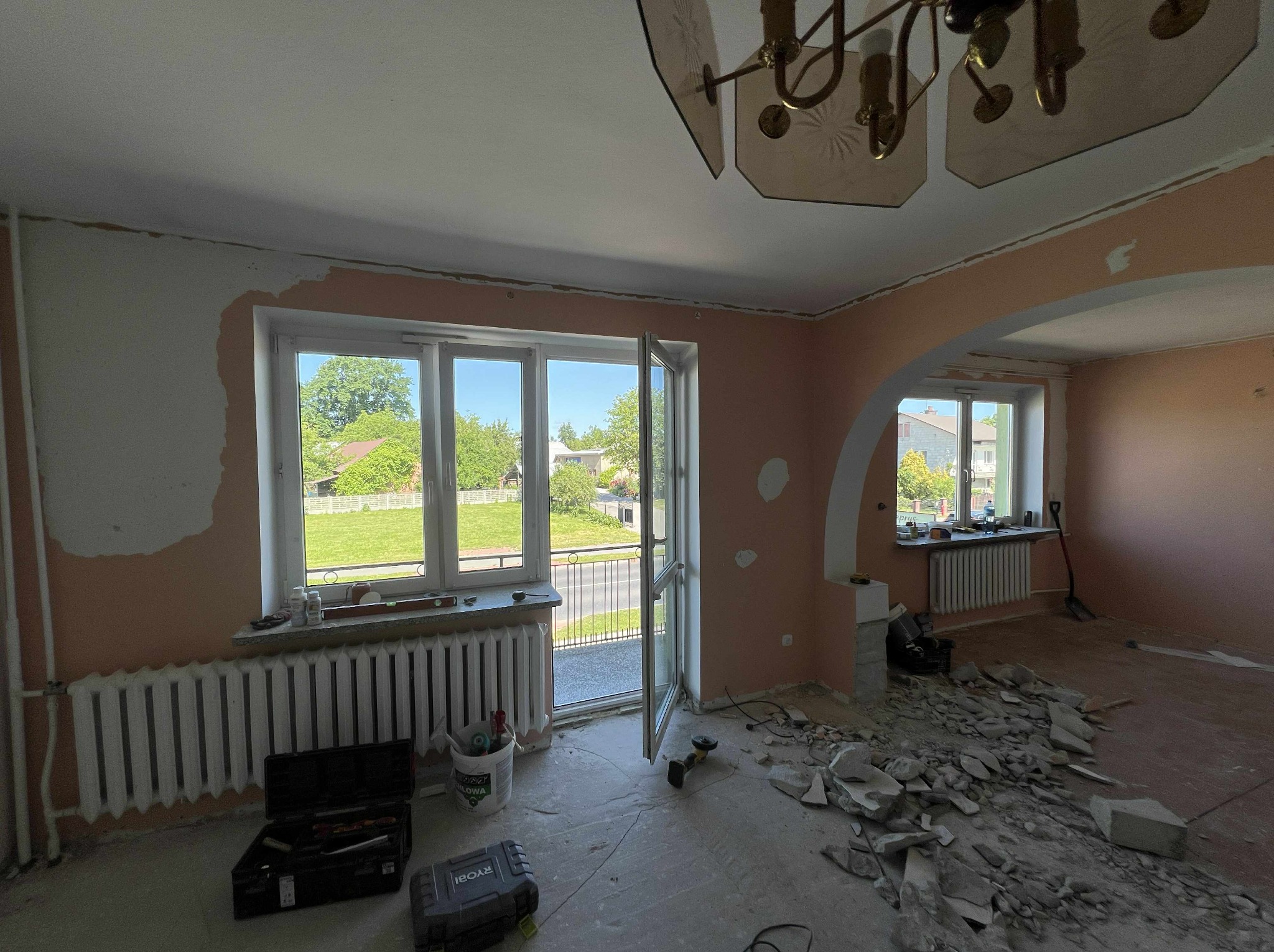 Demontaż podłogi w pokoju z oknami i kaloryferami. Fragmenty gruzu na podłodze, widoczne narzędzia i uszkodzone ściany. W tle zieleń za oknem.