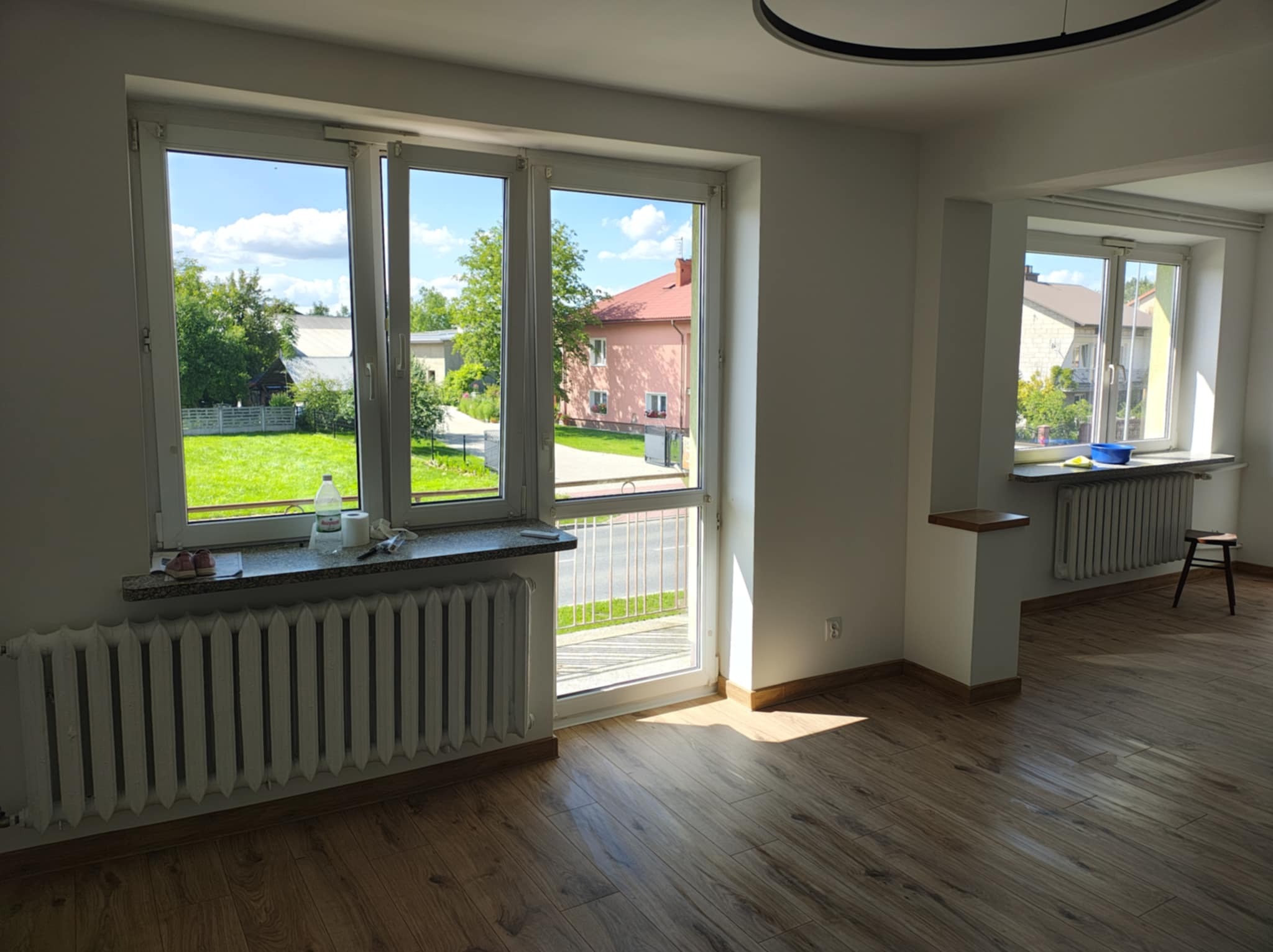 Jasne wnętrze po remoncie: drewniana podłoga, białe ściany, okna z widokiem na zieleń. Grzejniki pod oknami. Nowoczesna lampa sufitowa. Minimalistyczny wystrój.