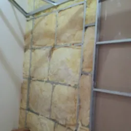 Wnętrze w trakcie wygłuszania: wełna mineralna między metalowymi profilami, częściowo zakryta płytą. Widoczne drewniane podpory. Narożnik pomieszczenia.