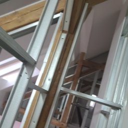 Jakub Michalski - Konstrukcja szkieletowa z profili stalowych i drewnianych belek podczas budowy poddasza, widok od dołu. Metalowe elementy tworzą geometryczny wzór.