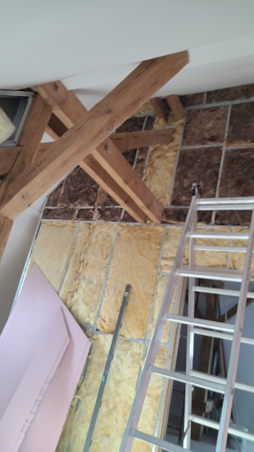 Izolacja poddasza z widocznymi belkami stropowymi, wełną mineralną i płytami izolacyjnymi. Aluminiowa drabina oparta o ścianę. Prace wykończeniowe.