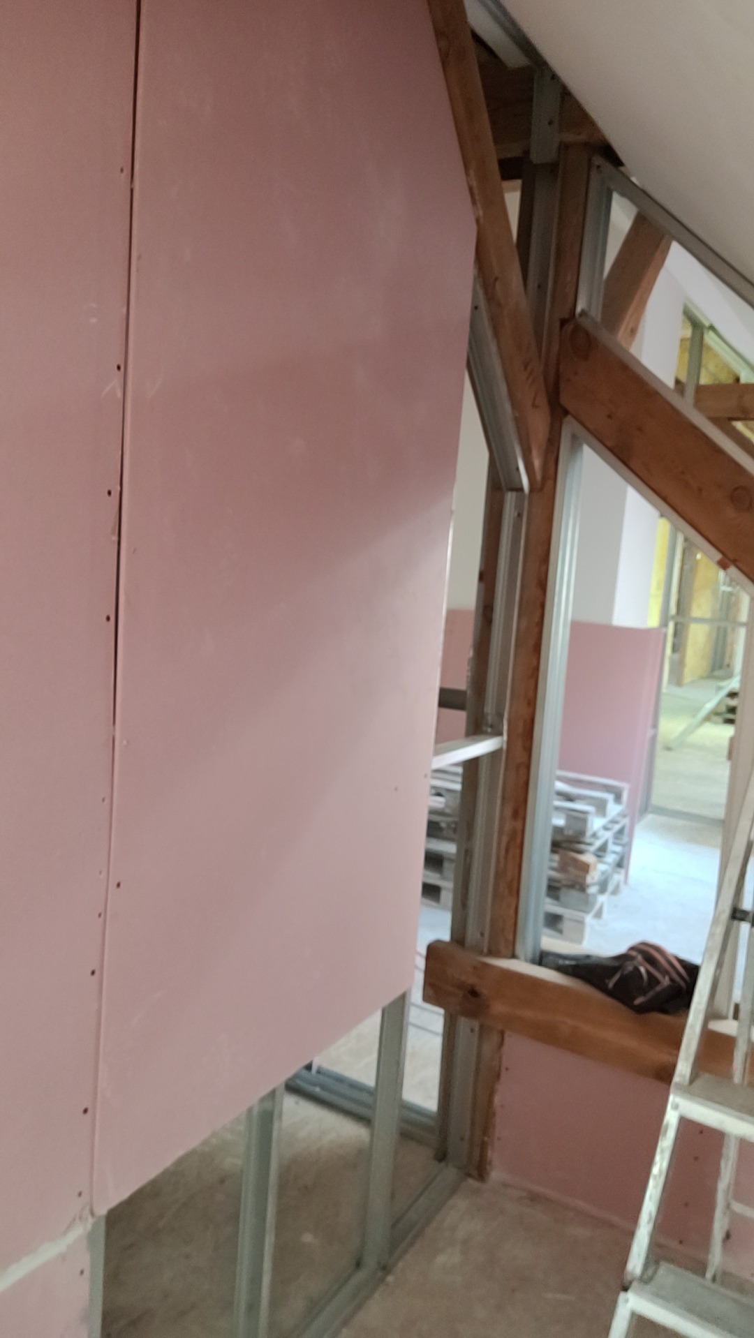 Montaż płyt gipsowo-kartonowych w poddaszu, widoczne metalowe profile i drewniane elementy konstrukcyjne. Fragment ściany pomalowany na różowo.