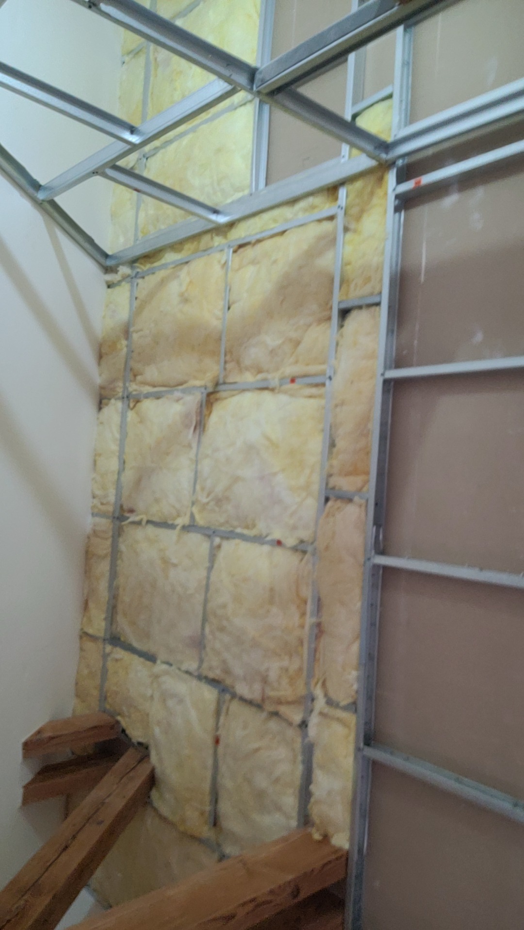 Wnętrze w trakcie wygłuszania: wełna mineralna między metalowymi profilami, częściowo zakryta płytą. Widoczne drewniane podpory. Narożnik pomieszczenia.