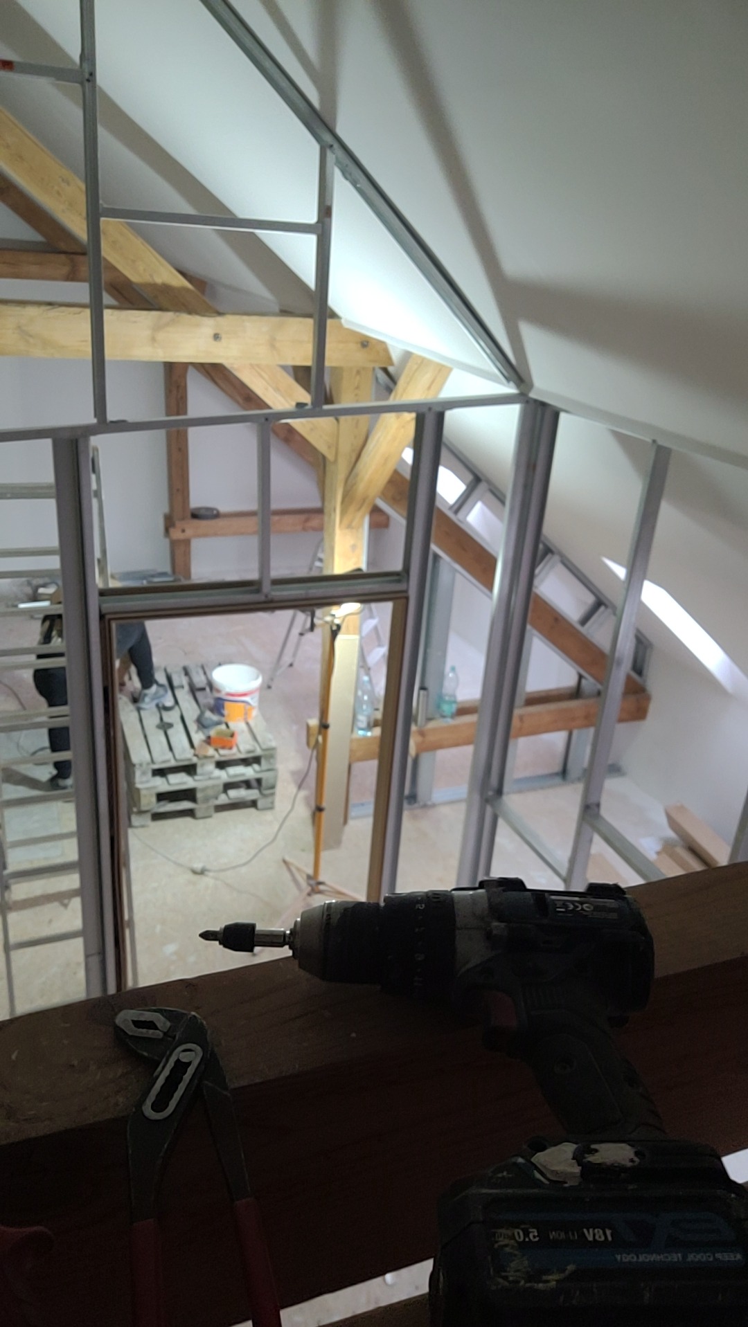 Montaż płyt na poddaszu: widoczne profile, narzędzia (wiertarka, kombinerki) oraz fragment drewnianej konstrukcji dachu. Prace wykończeniowe, remont.