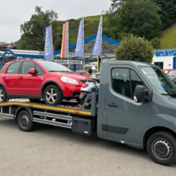 Szara laweta z dwoma czerwonymi autami Suzuki SX4 na platformie, na tle budynku warsztatu samochodowego i zielonego wzgórza. Flagi reklamowe w tle.