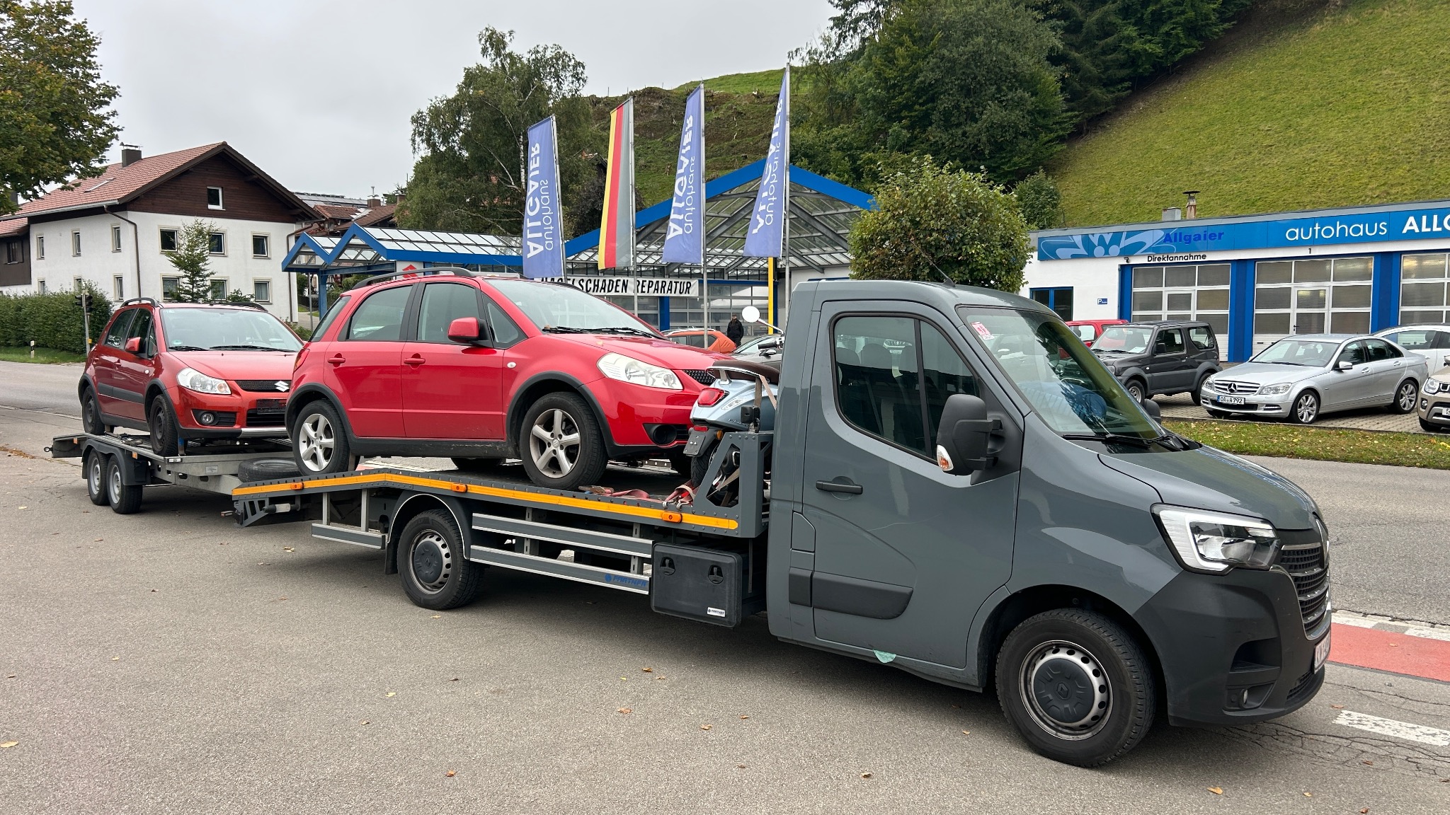 Szara laweta z dwoma czerwonymi autami Suzuki SX4 na platformie, na tle budynku warsztatu samochodowego i zielonego wzgórza. Flagi reklamowe w tle.