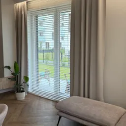 Jasne żaluzje poziome w oknie z widokiem na zieleń za budynkiem, zasłonięte beżowymi zasłonami w nowoczesnym salonie z drewnianą podłogą i rośliną doniczkową.