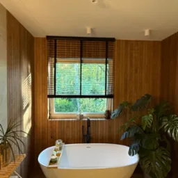 Elegancka łazienka z drewnianymi panelami, wolnostojącą wanną i żaluzjami drewnianymi w oknie z widokiem na zieleń. Minimalistyczny design i naturalne światło.