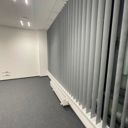 ROLETOWO MICHAŁ RYBCZYŃSKI - Szare żaluzje pionowe zamontowane w biurze, widoczne na tle ściany z gniazdkami elektrycznymi i szarą wykładziną. Perspektywa z dołu, oświetlenie sufitowe.