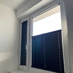 ROLETOWO MICHAŁ RYBCZYŃSKI - Ciemna plisa okienna na białym oknie PCV, częściowo zasłonięta, z widocznym mechanizmem sterowania. Jasne wnętrze, parapet z delikatnym odbiciem światła.