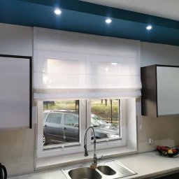 ROLETOWO MICHAŁ RYBCZYŃSKI - Jasna roleta rzymska, lekko opuszczona, w oknie kuchennym. Widok na zewnątrz, zlew i elementy wyposażenia kuchni. Błękitny sufit podwieszany z oświetleniem punktowym.