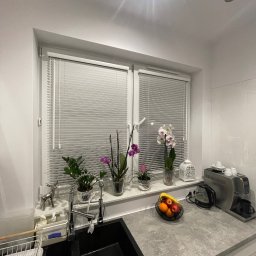 ROLETOWO MICHAŁ RYBCZYŃSKI - Kuchnia z oknem, na którym zamontowano białe, poziome żaluzje aluminiowe. Na parapecie kwiaty w doniczkach i dekoracje. Czarny zlewozmywak i szary blat.