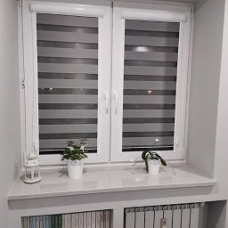 ROLETOWO MICHAŁ RYBCZYŃSKI - Białe okno z roletami dzień noc w szaro-białe pasy, z doniczkami na parapecie, wbudowane półki na książki i grzejnik pod oknem. Stonowana kolorystyka wnętrza.