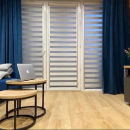 Eleganckie rolety dzień noc w nowoczesnym salonie z niebieskimi zasłonami i drewnianą podłogą. Laptop na stoliku kawowym dodaje wnętrzu przytulnego charakteru.