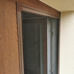 ROLETOWO MICHAŁ RYBCZYŃSKI - Zamknięta moskitiera w brązowej ramie, zamontowana w oknie o drewnianym wykończeniu, widoczna z bliska pod kątem.