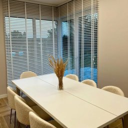 ROLETOWO MICHAŁ RYBCZYŃSKI - Jasne drewniane żaluzje poziome w nowoczesnej jadalni z białym stołem i beżowymi krzesłami. Dekoracyjny bukiet w wazonie na stole. Minimalistyczny design.