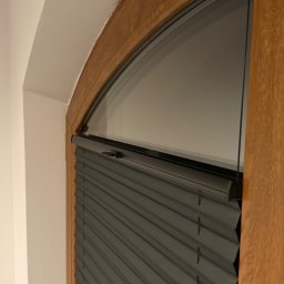 ROLETOWO MICHAŁ RYBCZYŃSKI - Szara plisa okienna zamontowana w drewnianej, łukowej ramie okiennej na tle białej ściany. Widoczne detale konstrukcji i faktura materiału.