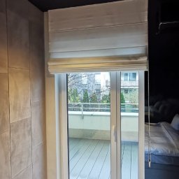 ROLETOWO MICHAŁ RYBCZYŃSKI - Elegancka roleta rzymska w jasnym odcieniu, zamontowana nad oknem balkonowym w nowoczesnej sypialni z ciemnym sufitem i jasną podłogą. Widok na balkon.