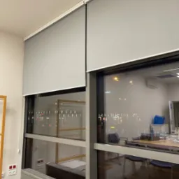 Dwie szare rolety wewnętrzne zamontowane w oknach biurowych. Widoczny mechanizm rolety i fragment wnętrza biura z widokiem na miasto nocą.