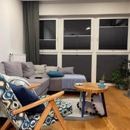 ROLETOWO MICHAŁ RYBCZYŃSKI - Ciemne plisy okienne w nowoczesnym salonie z szarą sofą i drewnianym stolikiem kawowym. Widok na okna z plisami w neutralnym wnętrzu.