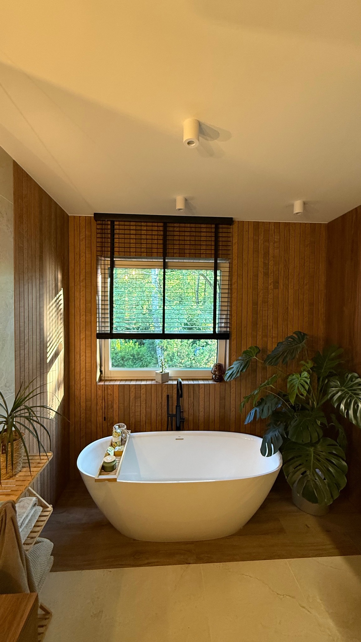 Elegancka łazienka z drewnianymi panelami, wolnostojącą wanną i żaluzjami drewnianymi w oknie z widokiem na zieleń. Minimalistyczny design i naturalne światło.