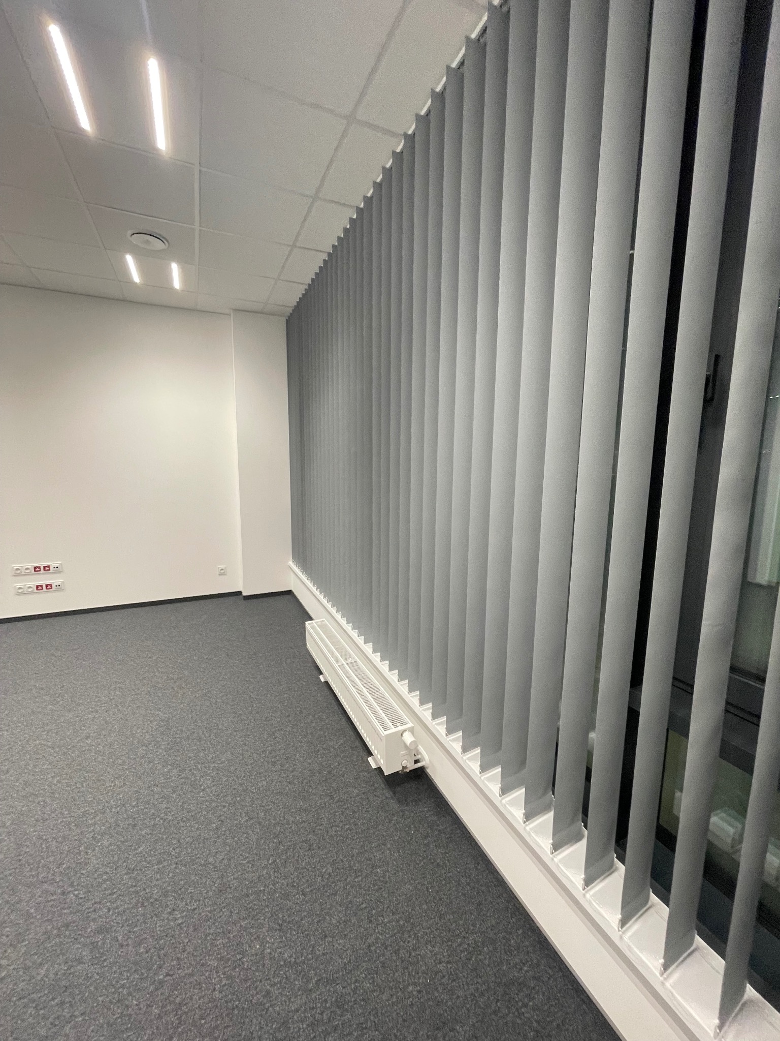 Szare żaluzje pionowe zamontowane w biurze, widoczne na tle ściany z gniazdkami elektrycznymi i szarą wykładziną. Perspektywa z dołu, oświetlenie sufitowe.