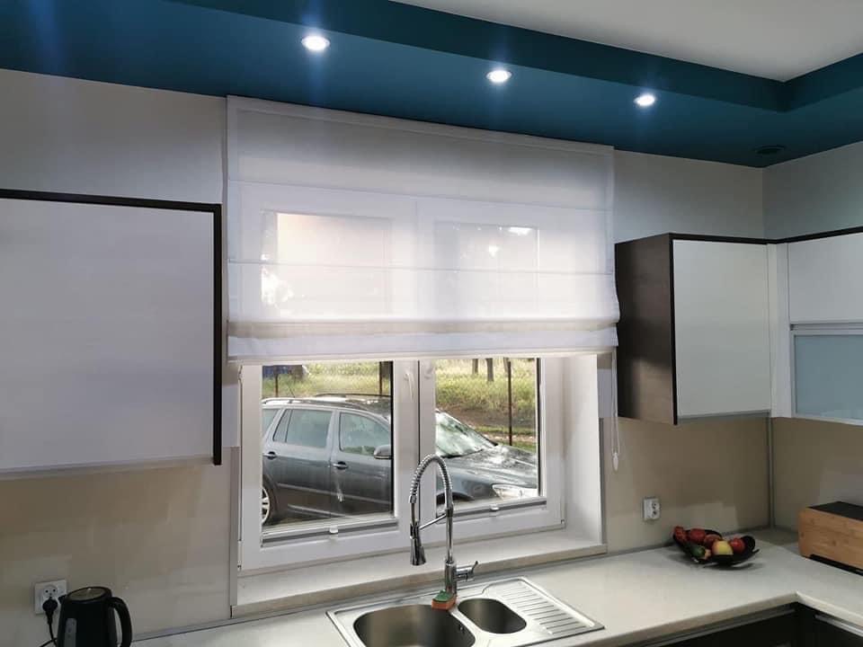 Jasna roleta rzymska, lekko opuszczona, w oknie kuchennym. Widok na zewnątrz, zlew i elementy wyposażenia kuchni. Błękitny sufit podwieszany z oświetleniem punktowym.