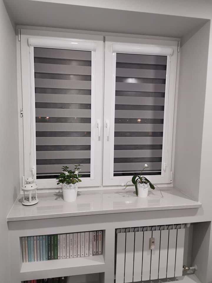 Białe okno z roletami dzień noc w szaro-białe pasy, z doniczkami na parapecie, wbudowane półki na książki i grzejnik pod oknem. Stonowana kolorystyka wnętrza.