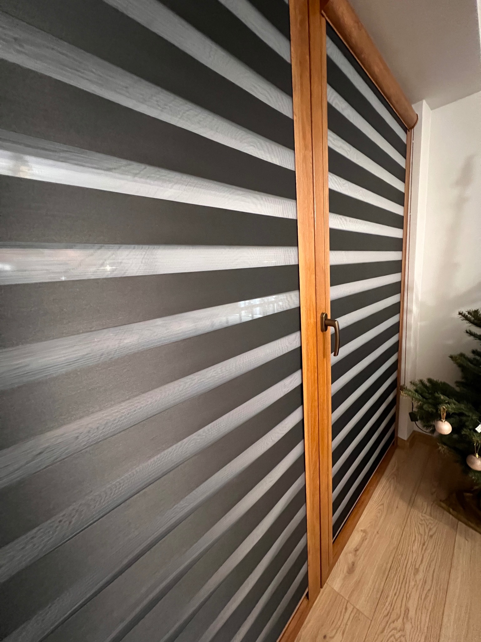 Zamknięte rolety dzień noc w drewnianej ramie okiennej, z widoczną częścią choinki w rogu, oświetlone naturalnym światłem.