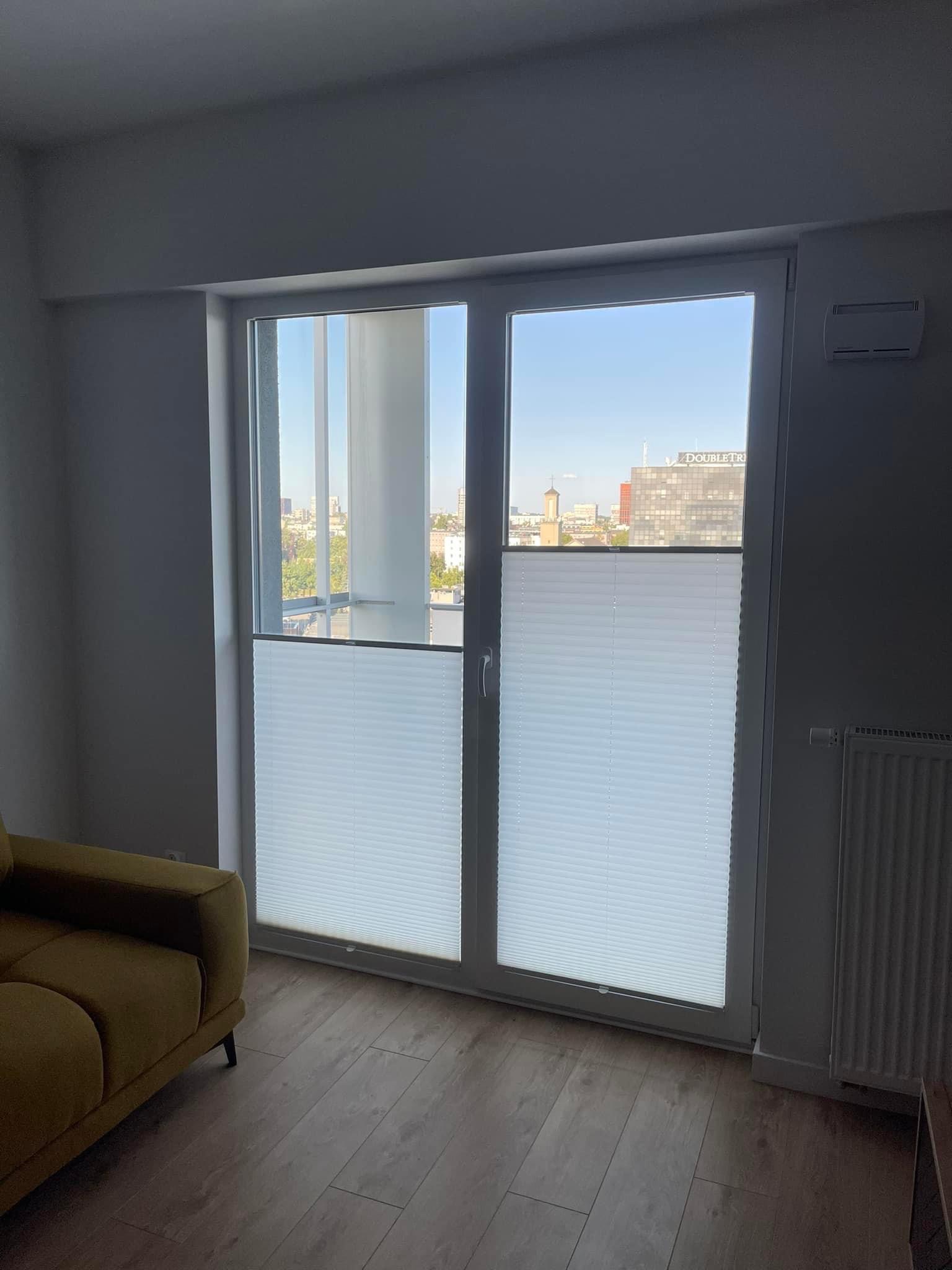 Dwuskrzydłowe okno z białymi plisami, widok na miasto. Częściowo widoczny żółty narożnik sofy i kaloryfer. Ujęcie z perspektywy wnętrza, parkiet na podłodze.