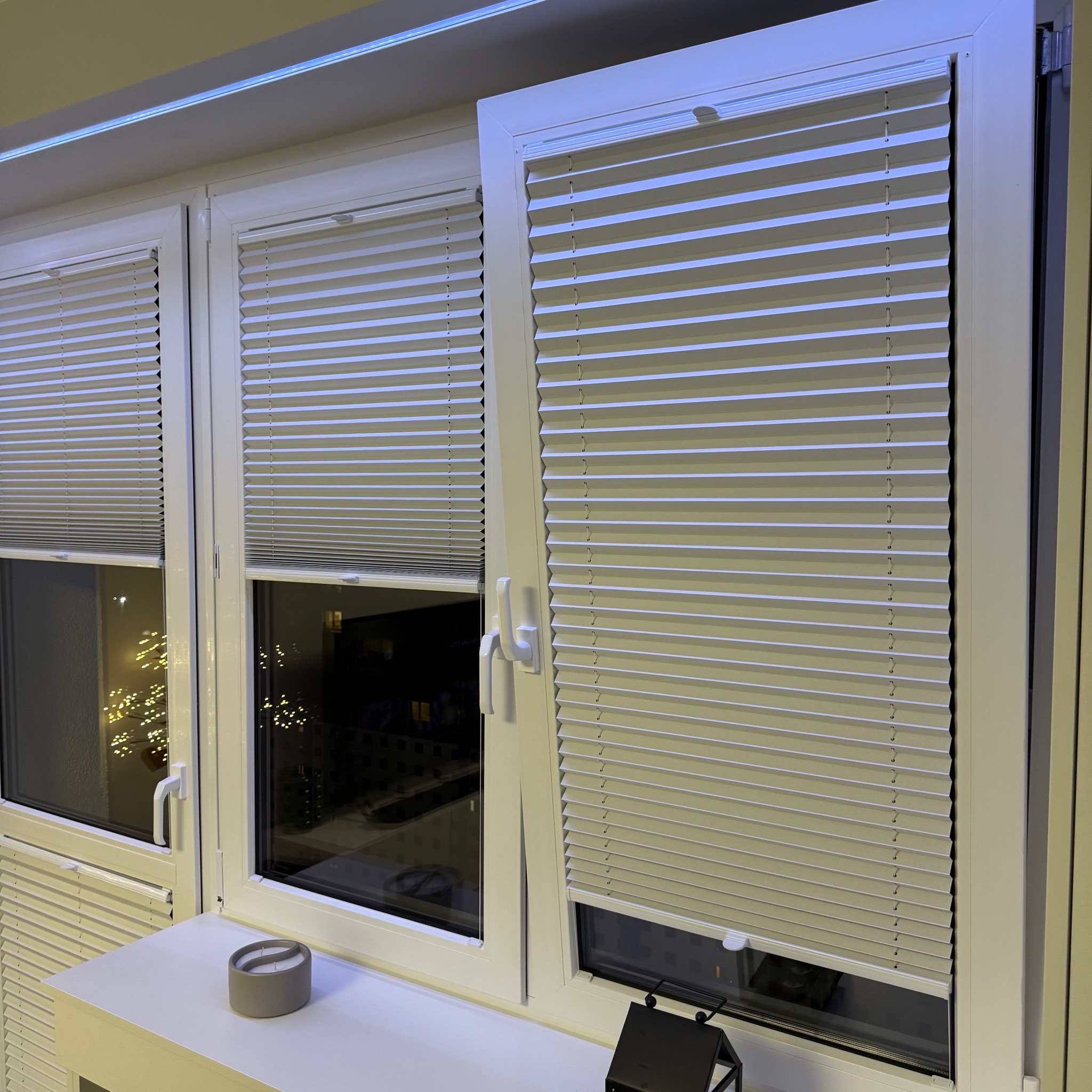 Trzy okna z białymi ramami, osłonięte plisowanymi roletami w kolorze ecru. Widok na nocne miasto i dekoracyjne światełka. Na parapecie świeczka i ozdobny lampion.